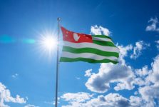 Abkhazian flag waving on pole, bright sun in the blue summer sky on the background